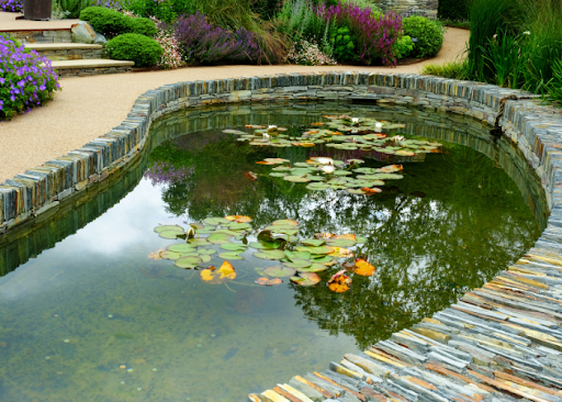 A small, clear ornamental pond with water lilies floating on the surface, bordered by neatly stacked stonework and surrounded by a landscaped garden with colorful flowers and greenery.