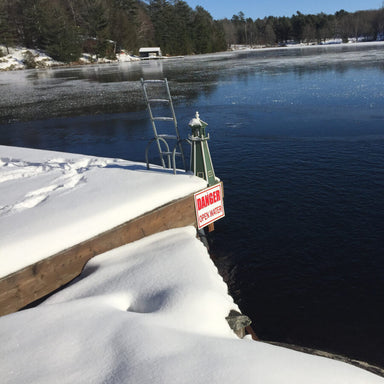 Frozen lake with snow-covered dock and warning sign, surrounded by trees and watel meted using the Ice Away De-Icer