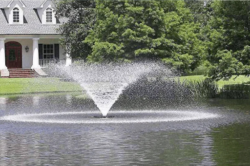 A pond fountain spraying water in front of a house and trees.