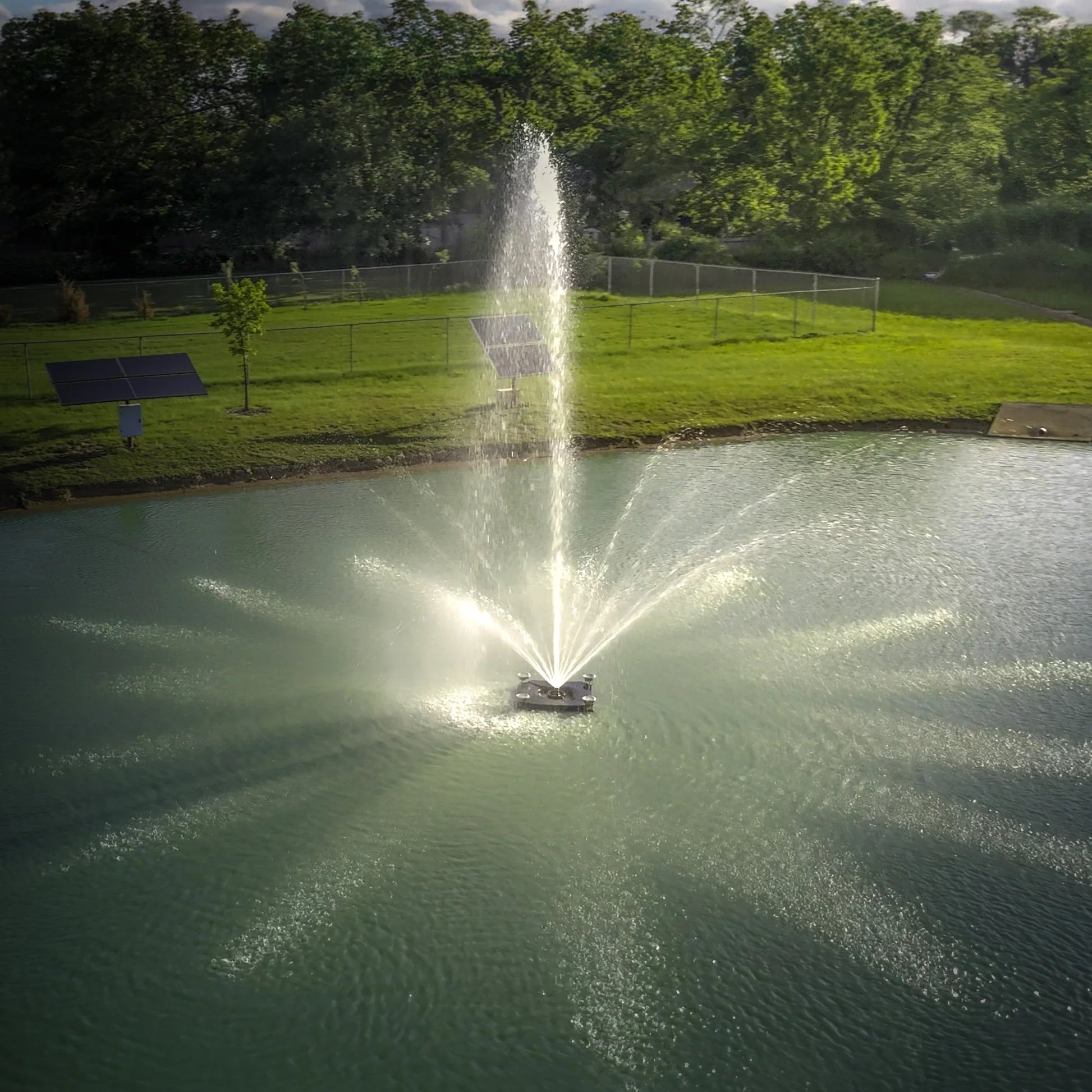 Fountain in a pond with solar panels, green grass and trees in the background