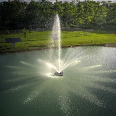 Fountain in a pond with solar panels, green grass and trees in the background