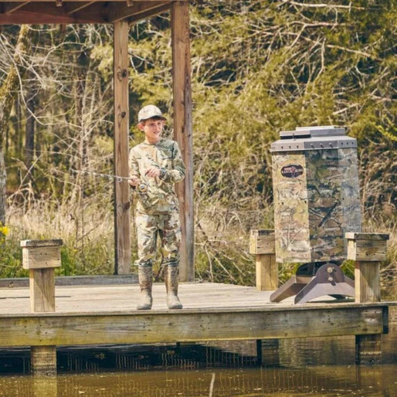 Person in camouflage standing on a wooden dock next to a Texas Avenger Fish Feeder
