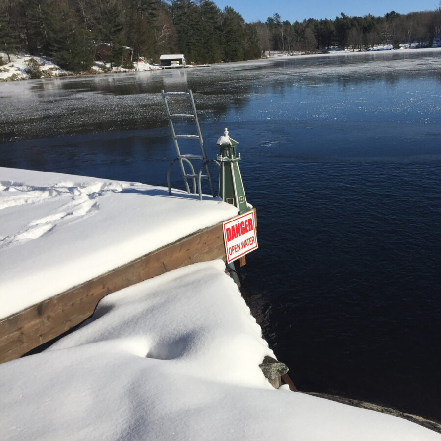 Frozen lake with snow-covered dock and warning sign, surrounded by trees and watel meted using the Ice Away De-Icer
